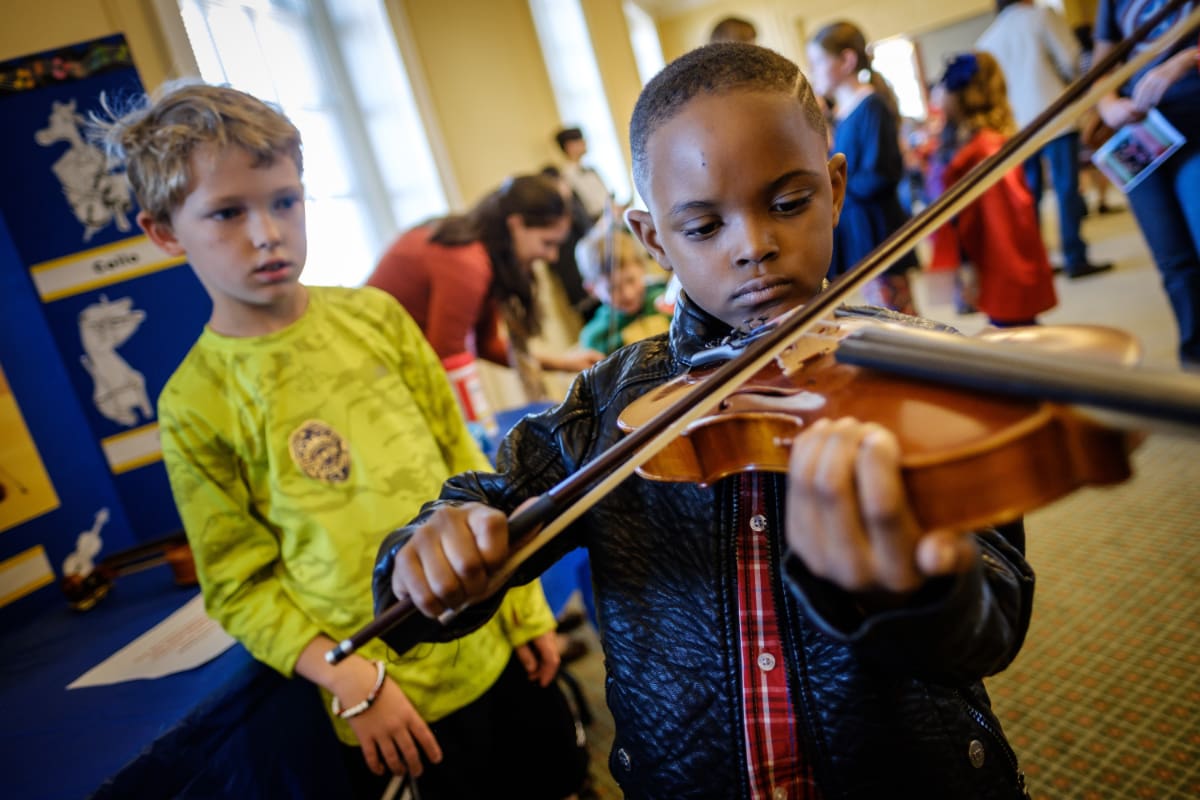 A child plays a violin at an Instrument Petting Zoo event while another child watches.