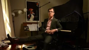 Ben Folds casually leaning against a pino with his feet up on a window sill.