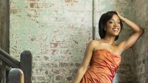 Violinist Kelly Hall-Tompkins in an orange dress against a white and light green brick wall.